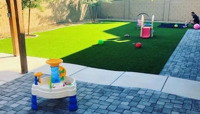 An Arizona back yard with artificial turf, pavers, and a block wall — a toddler's water table and slide in the foreground, a parent sitting with a baby in the shade.